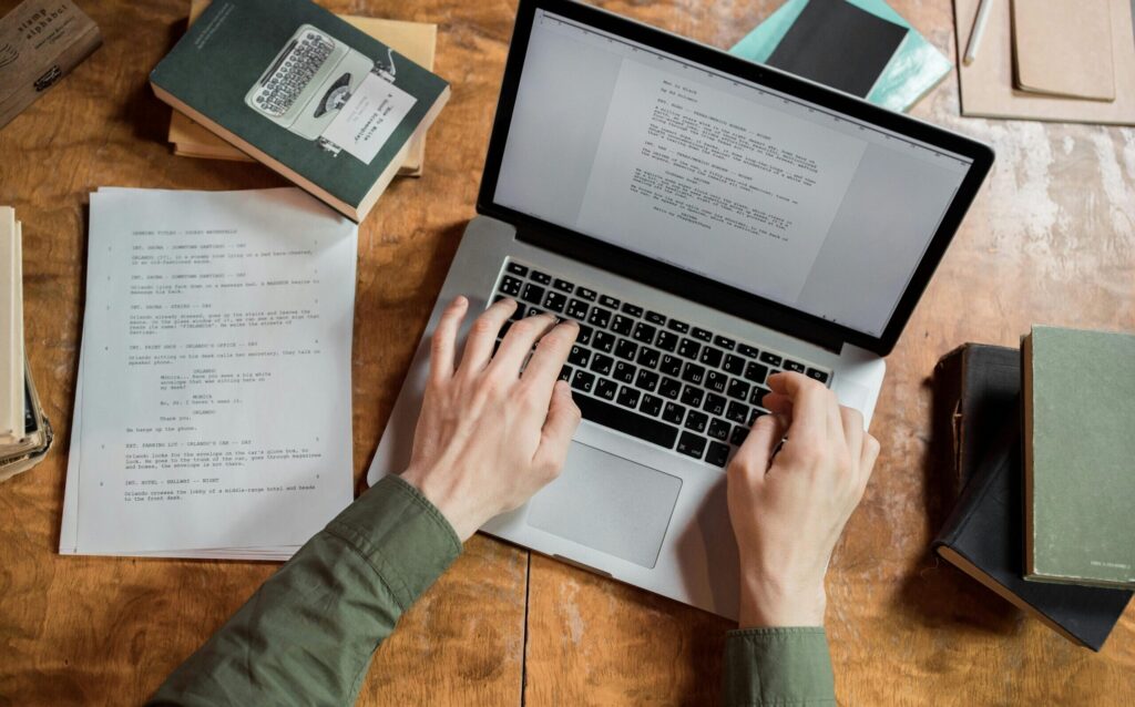 High-angle view of hands typing on a laptop surrounded by books and papers.